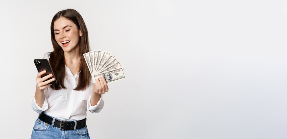 Retrato de uma mulher sorridente feliz usando um aplicativo de telefone movel segurando dinheiro em dinheiro de pe sobre um fundo branco